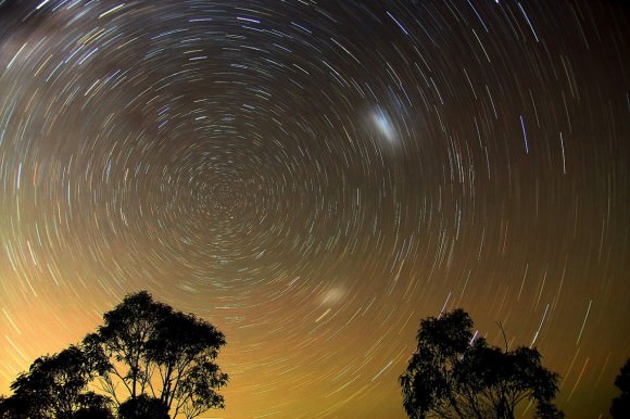 Stars trail around the dim southern pole star Sigma Octantis as seen from the southern hemisphere. The two smudges are the Large and Small Magellanic Clouds, companion galaxies of the Milky Way. Credit: Ted Dobosz