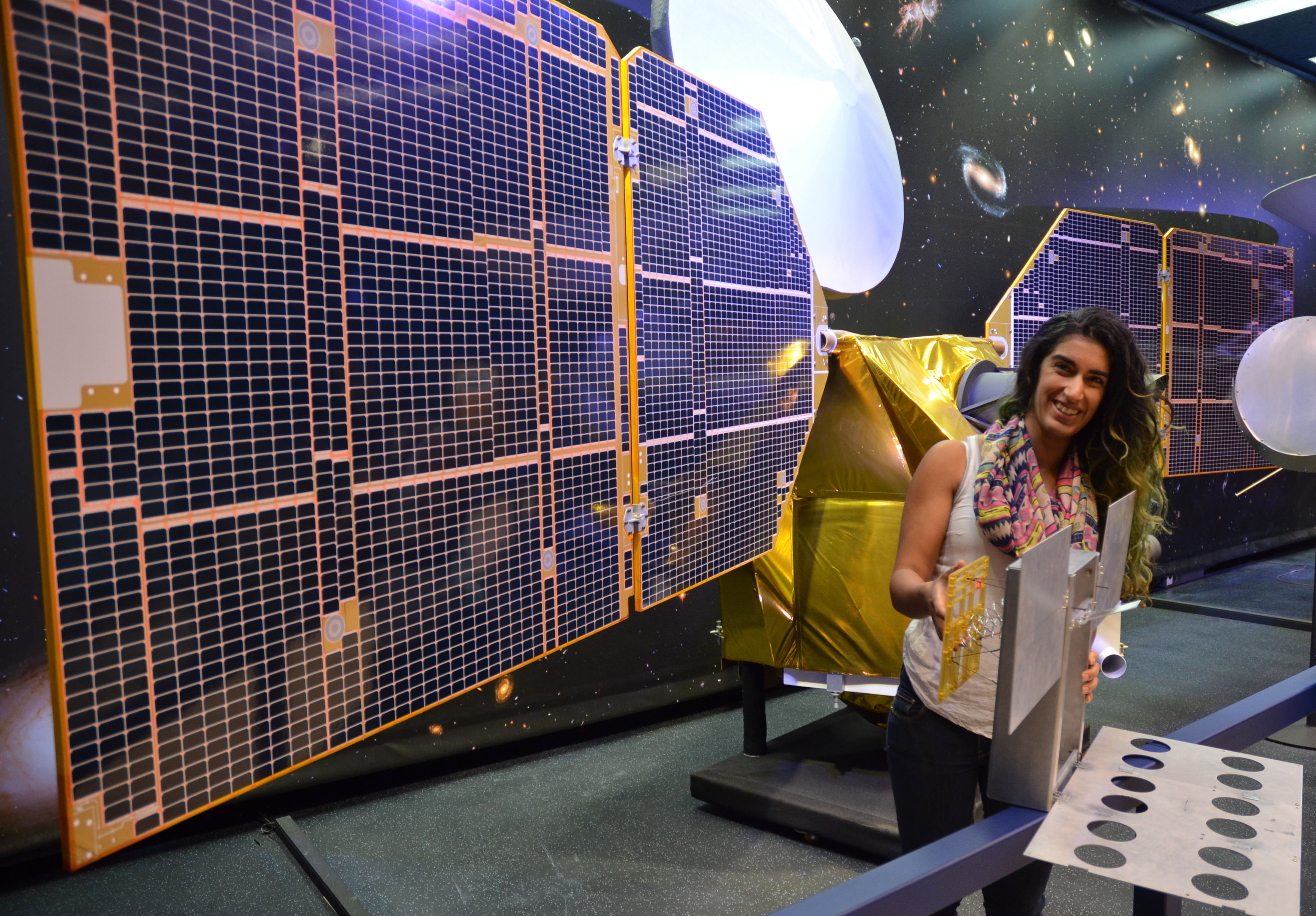 The full-scale mock-up of NASA's MarCO CubeSat held by Farah Alibay, a systems engineer for the technology demonstration, is dwarfed by the one-half-scale model of NASA's Mars Reconnaissance Orbiter behind her.  Credits: NASA/JPL-Caltech
