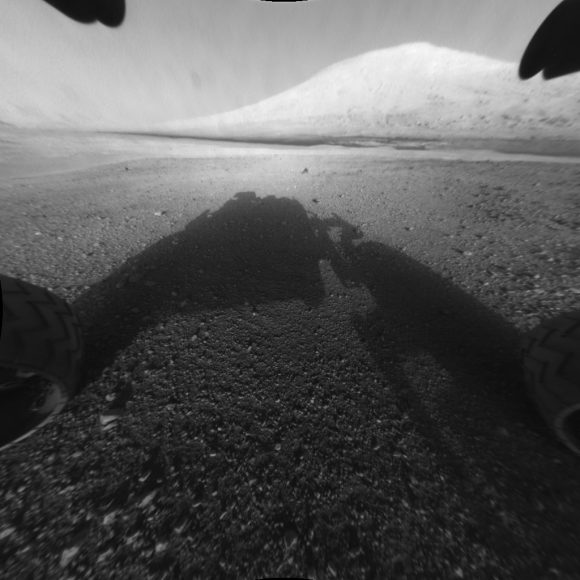 An image captured by the Curiosity rover shortly after it landed on the Red Planet on August 5, 2015, showing the rover's main science target, Mount Sharp. The rover's shadow can be seen in the foreground and the dark band beyond are dunes. Credit; NASA/JPL-Caltech.  