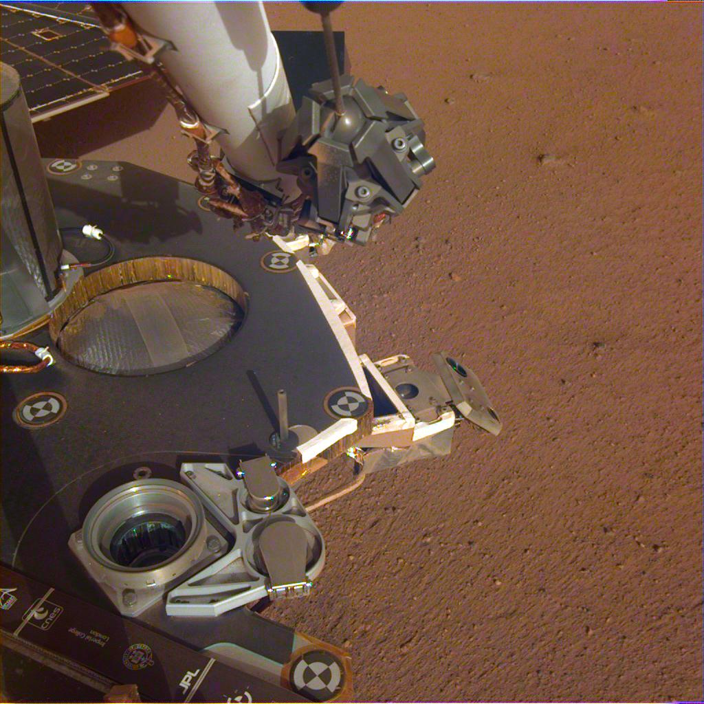 This is a partial view of InSight's deck. It shows the robotic arm and the stowed grapple. Sticking out in front of the deck is one of the boxy-looking attitude control thrusters used to guide the lander to the surface. In the middle of the image is the circular silver propellant tank inset into the lander's deck, and the cupholder-looking thing is one of the connections for InSight's aeroshell and parachute. Next to that is the UHF antenna which helps the lander communicate with Earth. The edge of one of the solar panels is visible on the far side of the deck. Image: NASA/JPL-Caltech