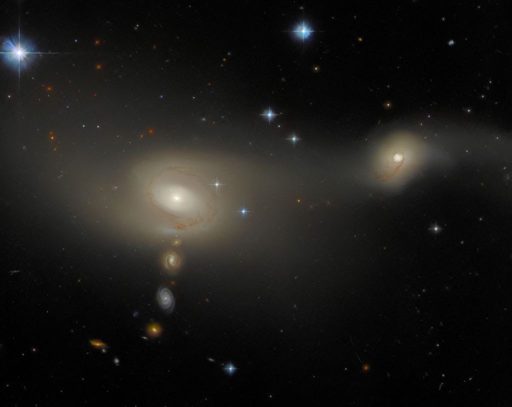 A pair of interacting galaxies, one smaller than the other. Each has a bright spot at the centre and two loosely-wound spiral arms, with threads of dark dust following the arms. They appear as a broad, soft glow in which individual stars can’t be seen. A number of bright stars and smaller, background galaxies can also be seen — three such galaxies lie in a vertical line below the right-hand galaxy of the pair.