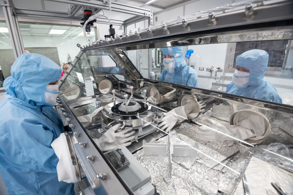 OSIRIS-REx astromaterials processors, from left, Rachel Funk, Julia Plummer, and Jannatul Ferdous, prepare to lift the top plate of the Touch-and-Go Sample Acquisition Mechanism (TAGSAM) head and pour the final portion of asteroid rocks and dust into sample trays below. Credit: NASA/Robert Markowitz